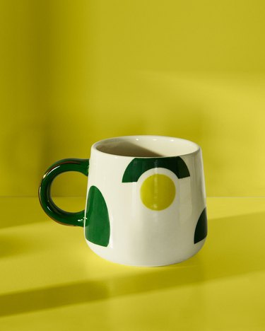 White mug with dark and light green geometric shapes representing an avocado. The mug is on a lime green background.