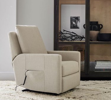 Tan reclining chair with a remote control in a side pocket on a tan carpet in front of white walls