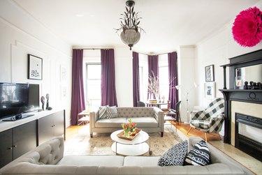 Photo of a room designed by AphroChic. The living room is wide and spacious, with two beige couches arranged in the middle. Navy and white pillows with various speckled patterns sit on the couches.  A flat screen TV sits on the white and chestnut bureau set up on the left wall, and a black and white mantle and fireplace is arranged on the right. The room features fun flourishes like a black and white gingham-patterned chair, and a fuschia loofah-like wall art.