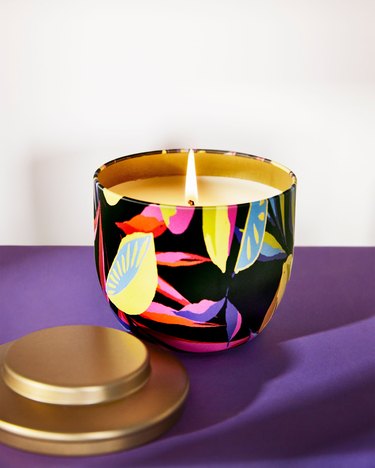 Black candle with bold colors resembling leaves and a gold lid sitting on a purple surface