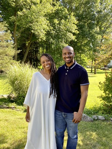 Photo of Jeanine Hays and Bryan Mason, a black woman and man, standing close together in a park. Jeanine is wearing a  flowy white kaftan and her torso-length brown dreadlocks are pulled to the right side of her face. Bryan is bald with a black mustache; he is wearing a navy blue collared skirt and light denim jeans. They both have wide, inviting smiles.