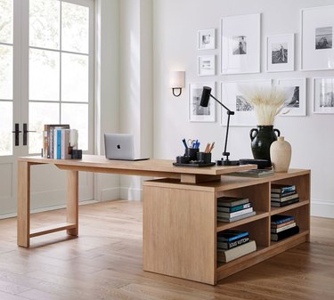 L-shaped desk topped with books, a laptop, and lamp, with books on the side in shelves. The wooden desk is on a wooden floor in front of windows and a gallery wall