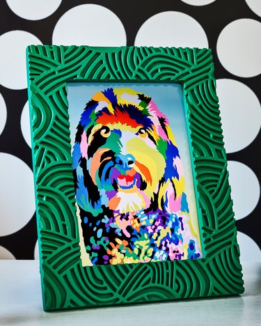 Colorful painting of a dog in a green and black frame. The frame sits on a black and white background.