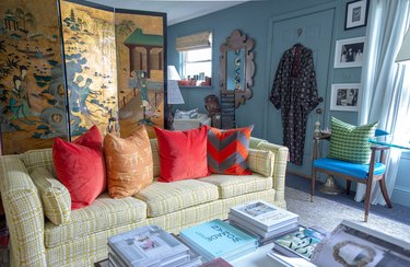 Photo of a living room designed by Dressing Room Interiors. The room is cozy and vibrant, with baby blue walls and two windows. There is a pale yellow couch with four pillows that are either red or orange with various designs, including grey zigzags and chalk-like sketches. Various books are arranged on a coffee table, obscuring its surface. A folding screen is arranged behind the couch, displaying  a golden scene of a river near a temple.