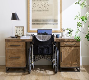 Wheelchair in front of a wooden desk. The desk has a coffee mug, clock and lamp. It sits in front of a white wall with a tan painting on it.