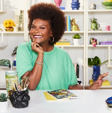 Tabitha Brown in a bright green dress sitting at a white desk with a water bottle, stationary, and a cup holder full of pens. Behind Brown is a white shelve with vases, books, and flowers.