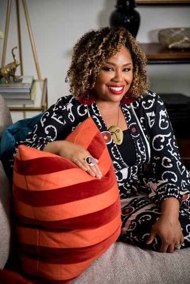 Photo of Ariene Bethea, a black woman with brown and blonde curly hair right above her shoulders. She is wearing a black and white dress that showcases a fun swirly pattern. Her gold necklace has an ovular face at the end, showing two eyes, eyebrows, a nose, and a mouth. She has her hand on a red and orange striped throw pillow.