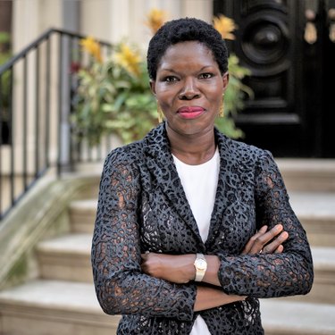 Photo of Nana Quagraine, a black woman with a black afro that is cut short. She is wearing a black and blue-grey blazer with toile-like embellishments over a crisp white shirt. She stands in front of a steps of a building, her arms crossed, a white and gold watch on her right wrist, and a warm smile on her face. She is wearing pink lipstick.