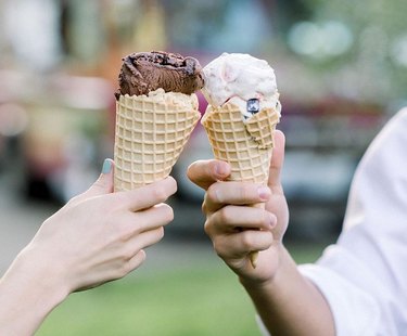 Two hands holding ice cream cones. One waffle cone has chocolate ice cream and the other waffle cone holds strawberry ice cream