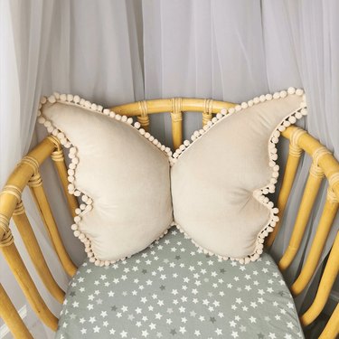 A beige butterfly-shaped pillow with pom poms on the edge. It is on a light wood chair with a light green seat cover that has white and dark green stars on it.