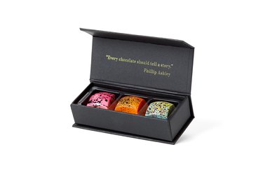 A box of three Philip Ashley Chocolates. Starting from the left, one is pink with brown chocolate splattered on top, one is orange with dots of chocolate, and the last is light blue with yellow and chocolate splotches. On the top of the box, it says "Every chocolate should tell a story."