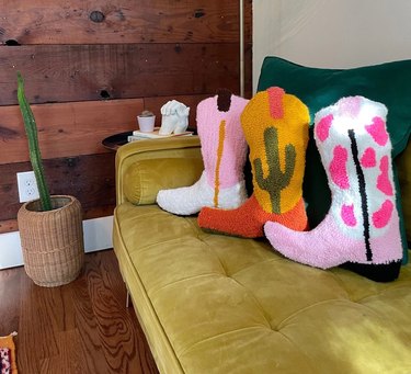 Three tufted cowboy boot pillows on a sage green couch. One is a pink and white bow pattern, another is yellow and orange and has a cactus on it, and the last is pink on top and white on the bottom with a vertical yellow stripe.