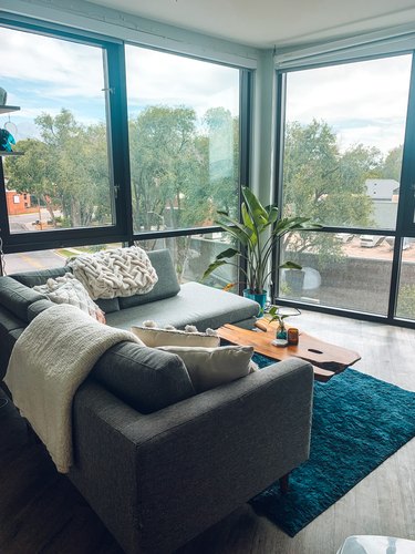 A modern gray sectional sofa with a chaise lounge in a living room apartment.  Floor to ceiling windows surround the space, with a live-edge wood coffee table and navy blue rug rounding out the space.
