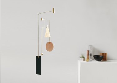 A mobile of geometric shapes made of brass, aluminum, and copper hangs in a white room with shelf in the background with small vases