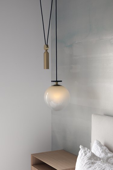 A black cord hangs from the ceiling in a corner with a small copper cylinder and warmly lit white globe in a room with a white wall opposite a sky patterned wall