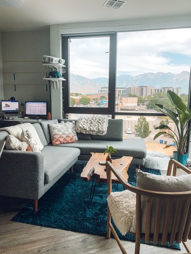 A modern gray sectional sofa with a chaise lounge in a living room apartment.  Floor to ceiling windows surround the space, with a live-edge wood coffee table and navy blue rug rounding out the space.