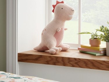 A pink tyrannosaurus rex pillow on a wood window ledge. The pillow also has dark pink spots and pink spikes on its head.