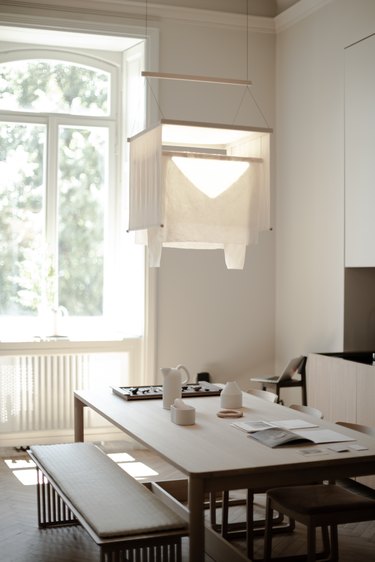 A cream colored fabric lamp hangs above a pale wood rectangular dining table with two matching rectangular benches near a window with sunshine coming through