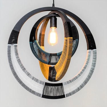 54kibo's Rope Pendant Light featuring three black wood circles, one inside the other. All the circles have open space at the bottom, where you can see the threading that has been wrapped around them. The smallest circle has blue thread, while the larger circles have yellow and white thread. There is a lightbulb at the center.