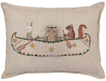 A beige pillow featuring a bunny, owl, and squirrel rowing a canoe in the center.