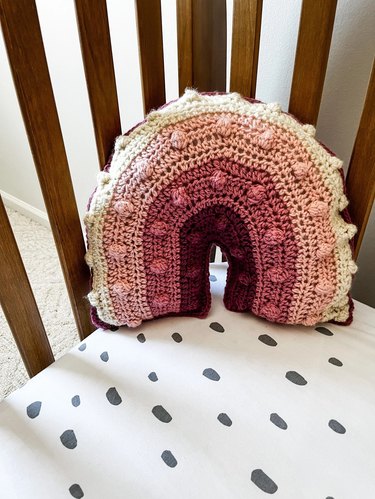 A crocheted rainbow pillow showing various shades of pink and white. It is show in a wood crib with a white sheet that has grey dots on it.