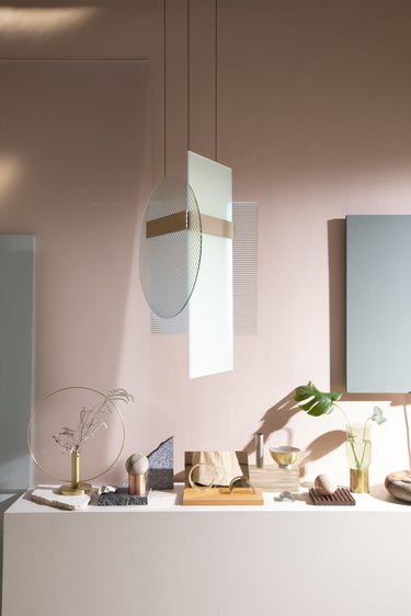 Pink room with shelf of different colored and shaped objects on it, a hanging lamp above it made of geometric glass shapes