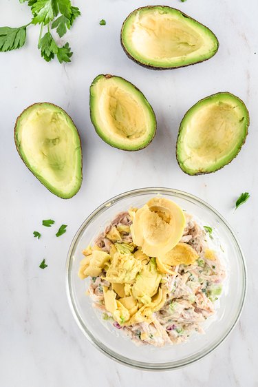 Four avocados with the pit scooped out. The bowl of shredded chick and the mayo mixture with mashed avocado inside it.
