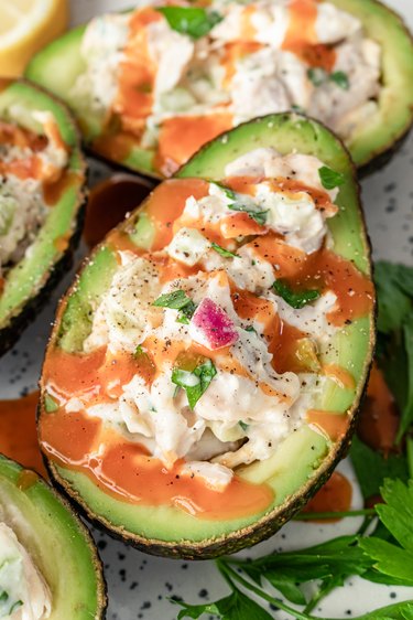 A close-up of an avocado filled with chicken salad. On top, there is a drizzle of hot sauce. In the background, you can see more avocados stuffed with chicken salad and part of a lemon.