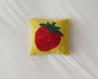 A mini square yellow pillow with a strawberry printed in the center. The strawberry has a red fruit dotted with medium-green lines. The stem and leaves at the top are also a medium green.