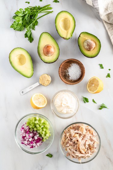 All the different ingredients laid out on a marble counter. The shredded chicken, chopped red onion and celery, and mayonnaise are in glasses containers. Dijon mustard is in a silver measuring spoon. Salt is in a wood bowl. There are also halved avocados and lemon on the counter with a sprig of cilantro.