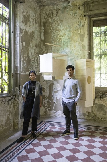 A dark-haired woman and man stand next to two hanging cream-colored fabric lamps in the corner of a high-ceiling room with chipped-paint walls and mauve and white diamond patterned floor