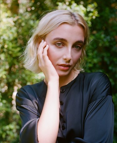 Emma Chamberlain, a white woman with white-blonde hair an inch above her shoulders. She has blue eyes, her right hand on her face, and is wearing a black button-up shirt and gold hoop earrings.
