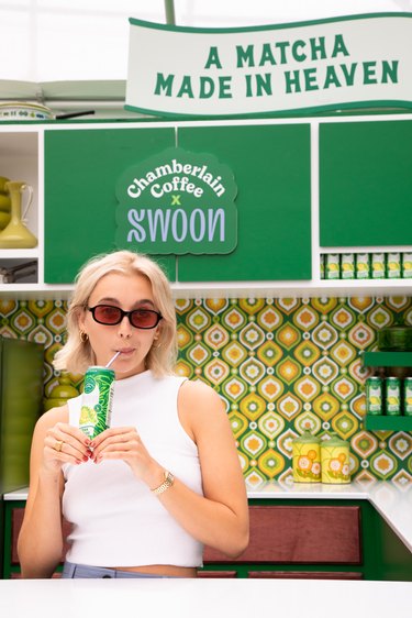 Emma Chamberlain drinking a can of Swoon Matcha Lemonade behind a white counter. In the back, there is '70s-inspired green and yellow wallpaper. You can also see the signs "Chamberlain Coffee x Swoon" and "A Matcha Made In Heaven." Emma has on black sunglasses with brown-tinted lenses and a white tank top.