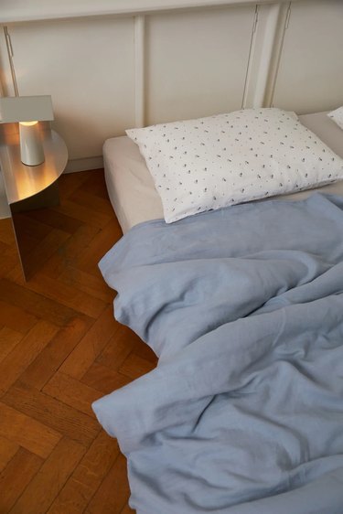 White floral pillow and a light blue duvet on a mattress on the hardwood floor.