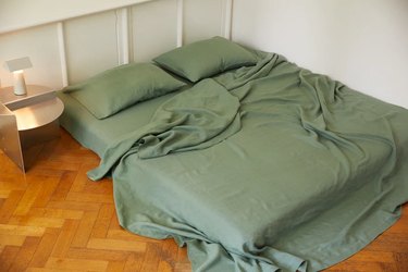 Green bedding on a mattress that sits on a hardwood floor. There is a small side table with a lamp and white walls.