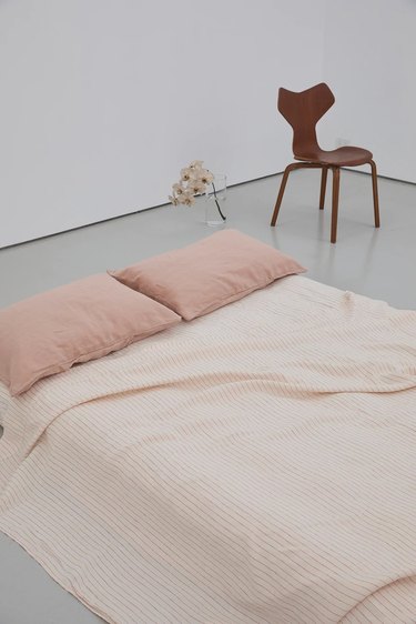 Clay pink sheets and blanket on a mattress on the floor. Behind the bedding is a white wall, a wooden chair, and pink flowers.