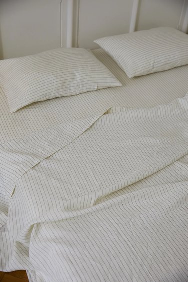 White bedding with light stripes on a mattress and sits on a floor in front of a white wall.