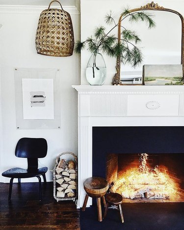 Fireplace with fairy lights, styled with an ornate gold mirror, vase with pine clipping, woven light, modern black side chair and wooden stools