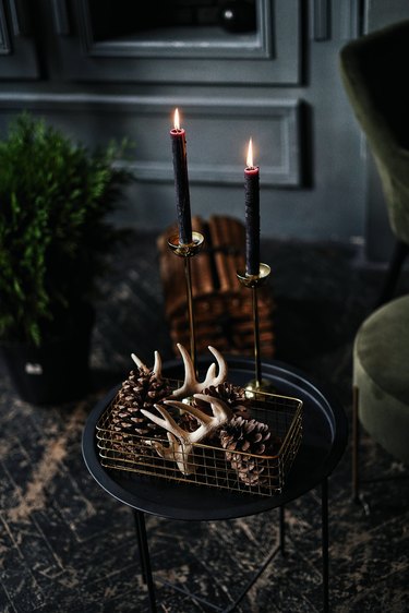 Antlers and pinecones sit in a metal basket atop a dark side table, with two candles glowing behind them.