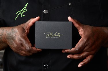Philip Ashley Rix holding a black closed box of his chocolates in between his two hands. On the box, his name is written in gold script. Above the box on the left, you can see an A written in script on Rix's black chef's jacket.