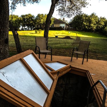 A door in a wooden deck leading down to a cavern. There are two outdoor chairs in the background and grass.