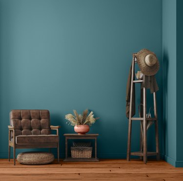 The teal Vining Ivy paint on a wall behind a brown armchair, side table topped with a fern plant, and a wood coat rack with hats on it.