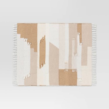 A patchwork throw blanket showing different brown, white, and beige geometric shapes. There is white fringe around the edge of the blanket.