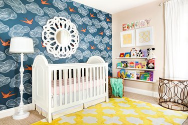 Baby's room with white crib, peacock blue/orange bird wallpaper and bright yellow patterned rug