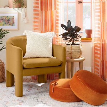 A mustard armchair with a curbed back in a living room by a window. There are two orange floor cushions on the floor, a white throw pillow on the chair, and orange striped curtains on the window.