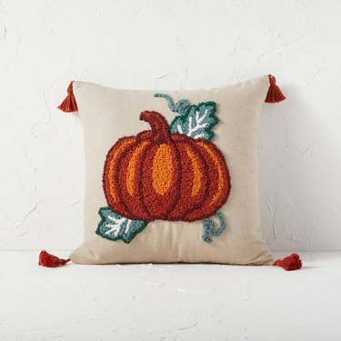 A beige throw pillow with a orange punch-needled pumpkin and green leaves on it. There is an orange tassel at each corner of the pillow.