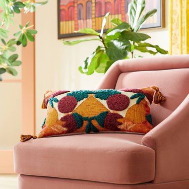 A lumbar throw pillow on a pink fabric armchair. The pillow features abstract green, yellow, and dark red shapes meant to convey pomegranates.