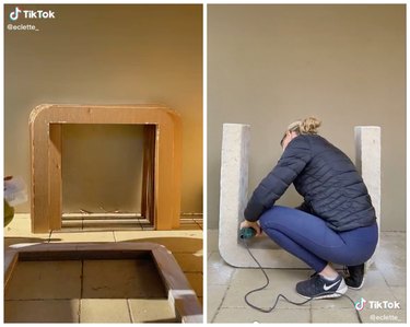 On the left is stack of cardboard shaped like a table leaning against a wall. On the right is a woman in a navy blue jacket sanding down a table.