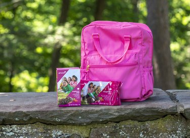 Two boxes of Raspberry Rally Girl Scout Cookies in front of a pink backpack resting on a ledge with trees in the background
