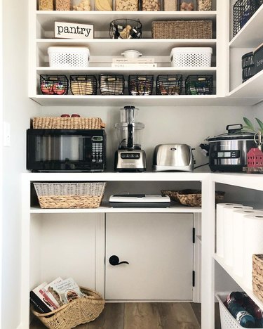 A pantry with white shelving and a small square door at the bottom with a black door knob. There are various appliances and foods on the shelves.